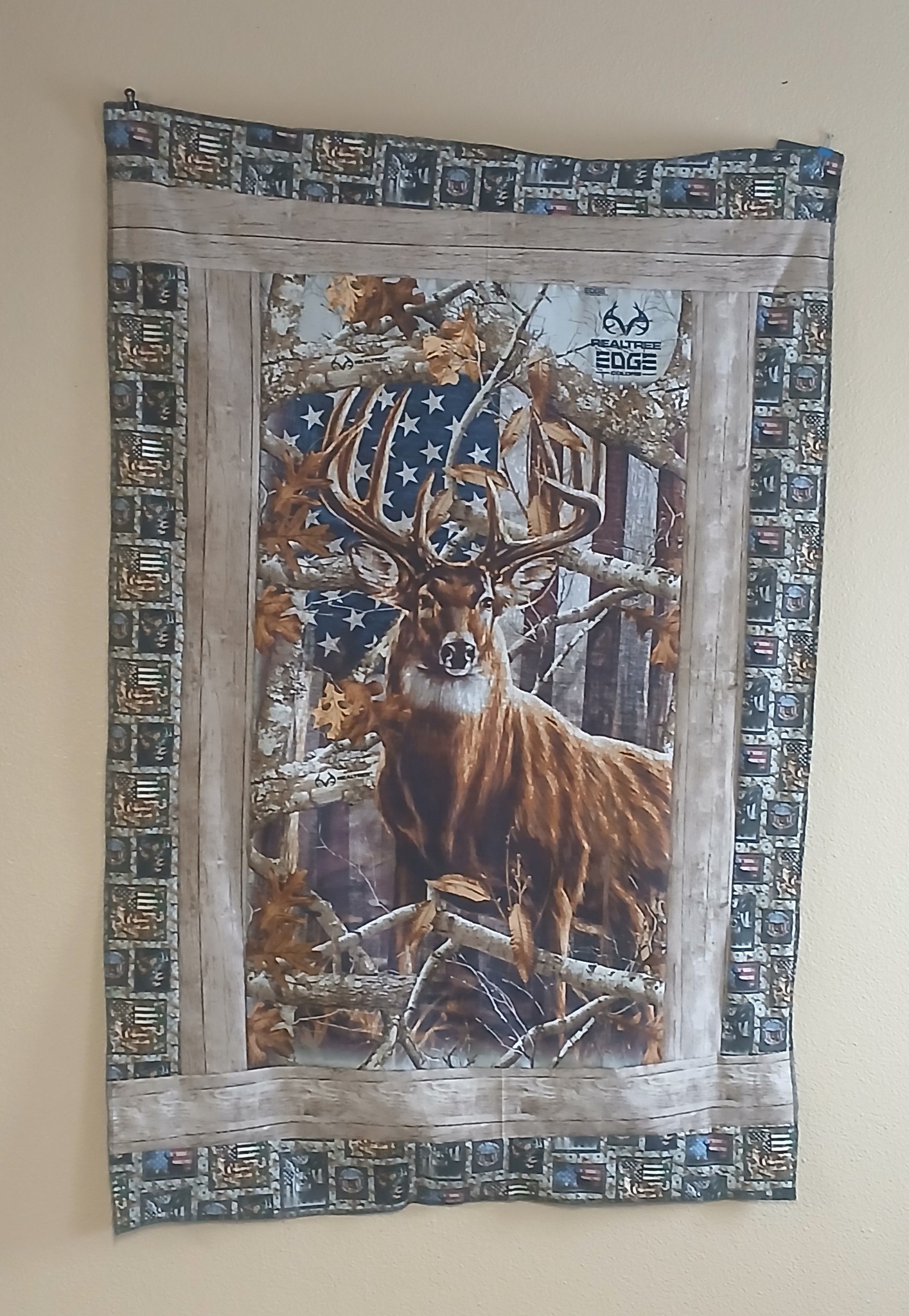 A fabric wall hanging featuring a majestic deer in a forest setting with an American flag background, framed with a rustic wooden border and additional decorative elements.